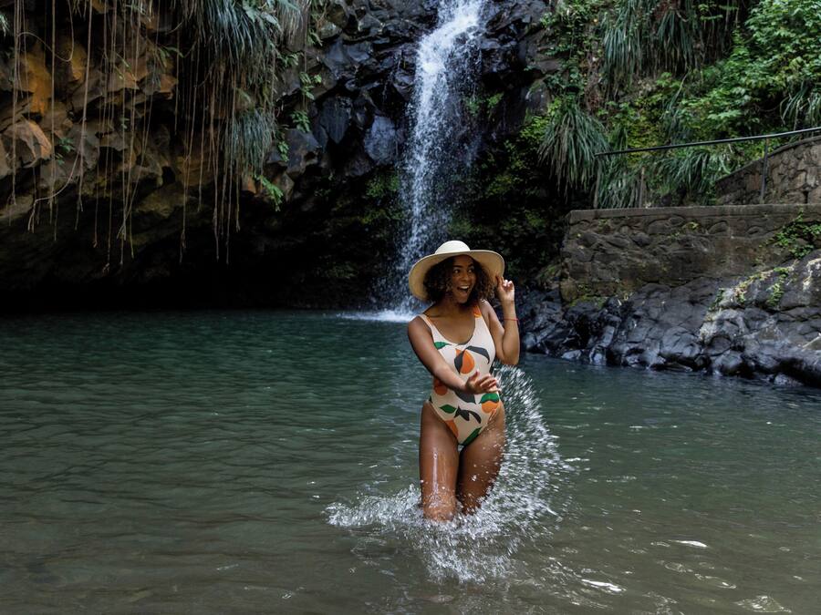 Una persona en traje de baño y sombrero, de pie en el agua, con una cascada al fondo.
