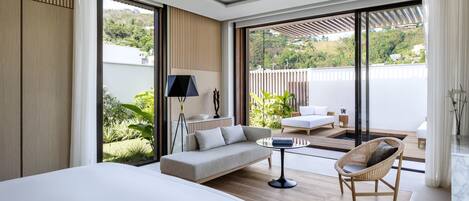 Une chambre moderne avec un grand lit, un canapé, un fauteuil et une petite table. Elle dispose d’un balcon offrant une vue sur la verdure et les bâtiments.