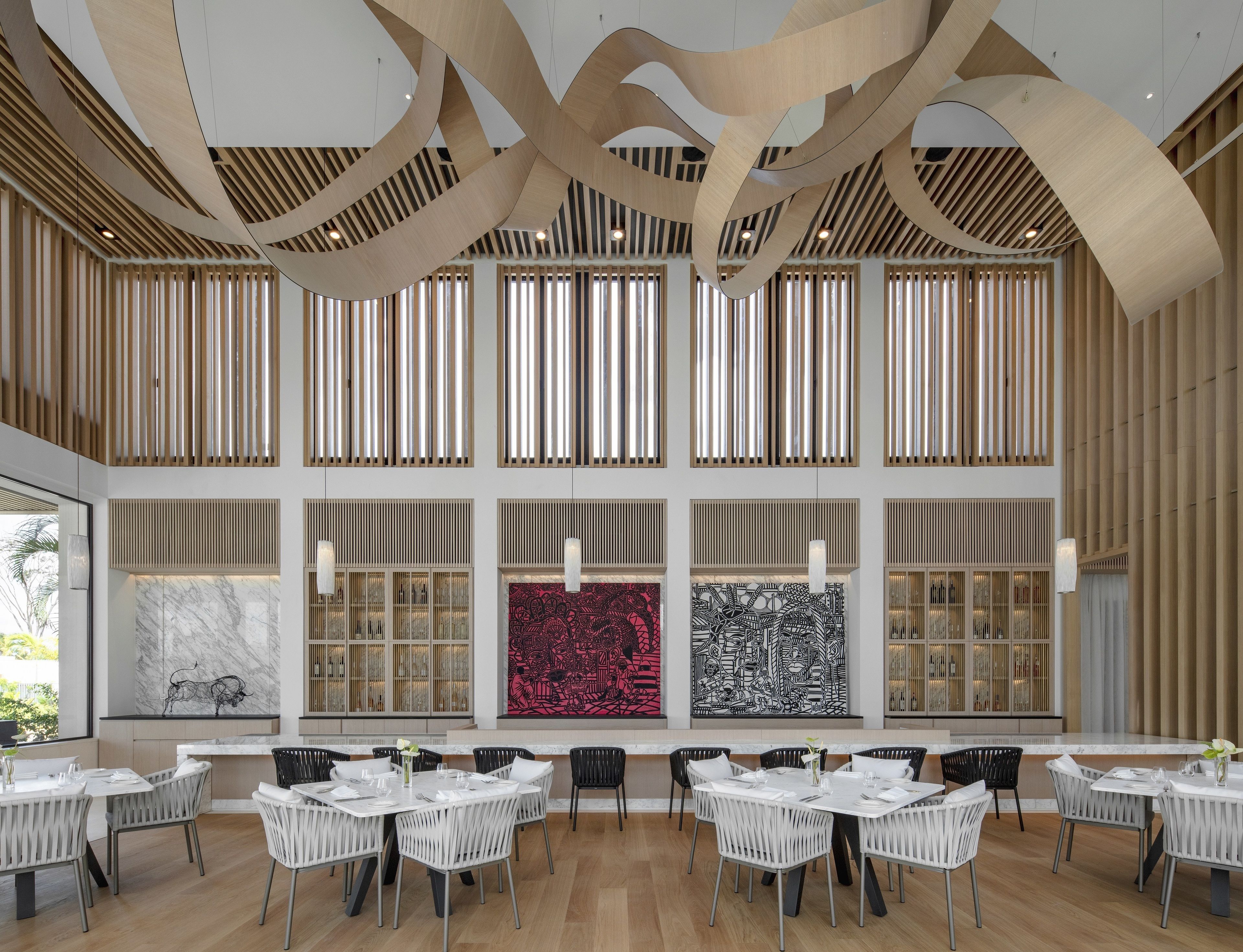 Un comedor moderno con sillas y mesas blancas, listones de madera en el techo y obras de arte en las paredes.