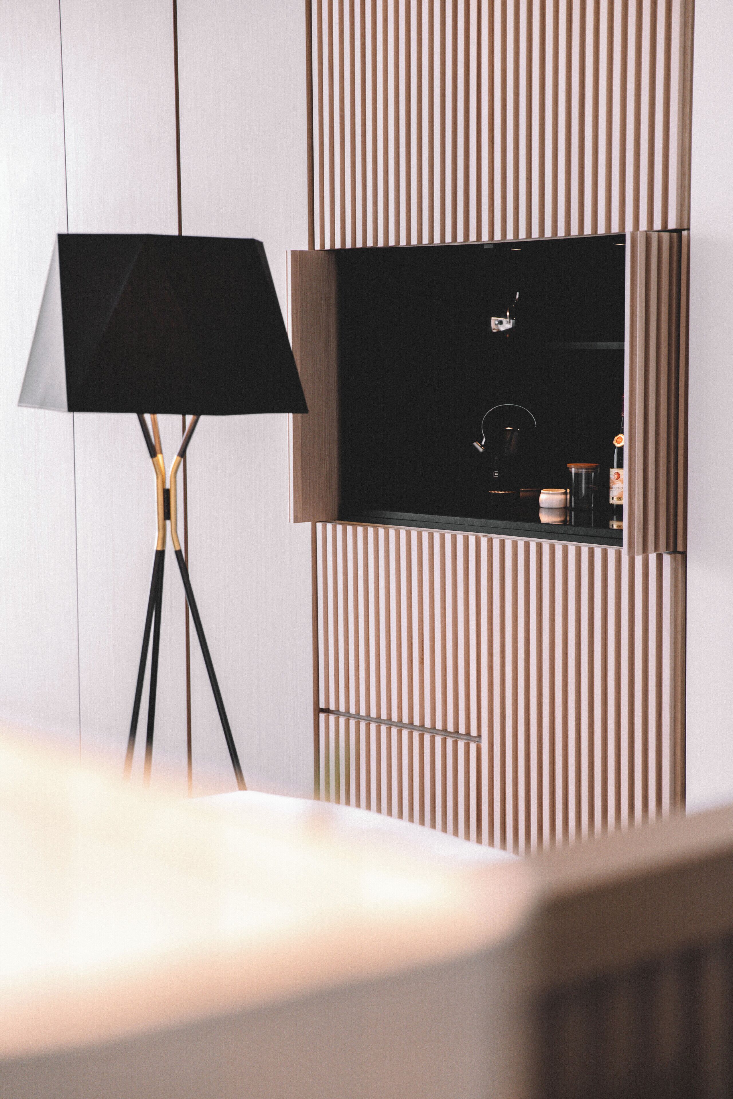Una habitación moderna con una lámpara de pie, televisor montado y una zona de cocina equipada con hervidor y cafetera.