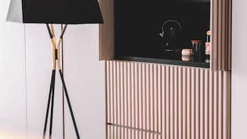 Una habitación moderna con una lámpara de pie, televisor montado y una zona de cocina equipada con hervidor y cafetera.