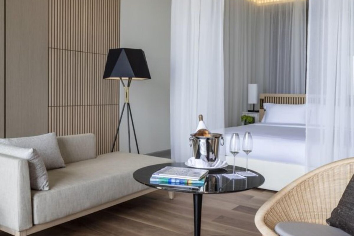 Une chambre d’hôtel moderne avec un canapé, une table ronde agrémentée de livres et de verres, une lampe et un lit à baldaquin.
