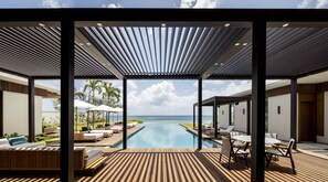 A modern outdoor pool area with wooden decking, lounge chairs, and umbrellas, overlooking a clear blue sea. - Silversands Grand Anse (St. George's)