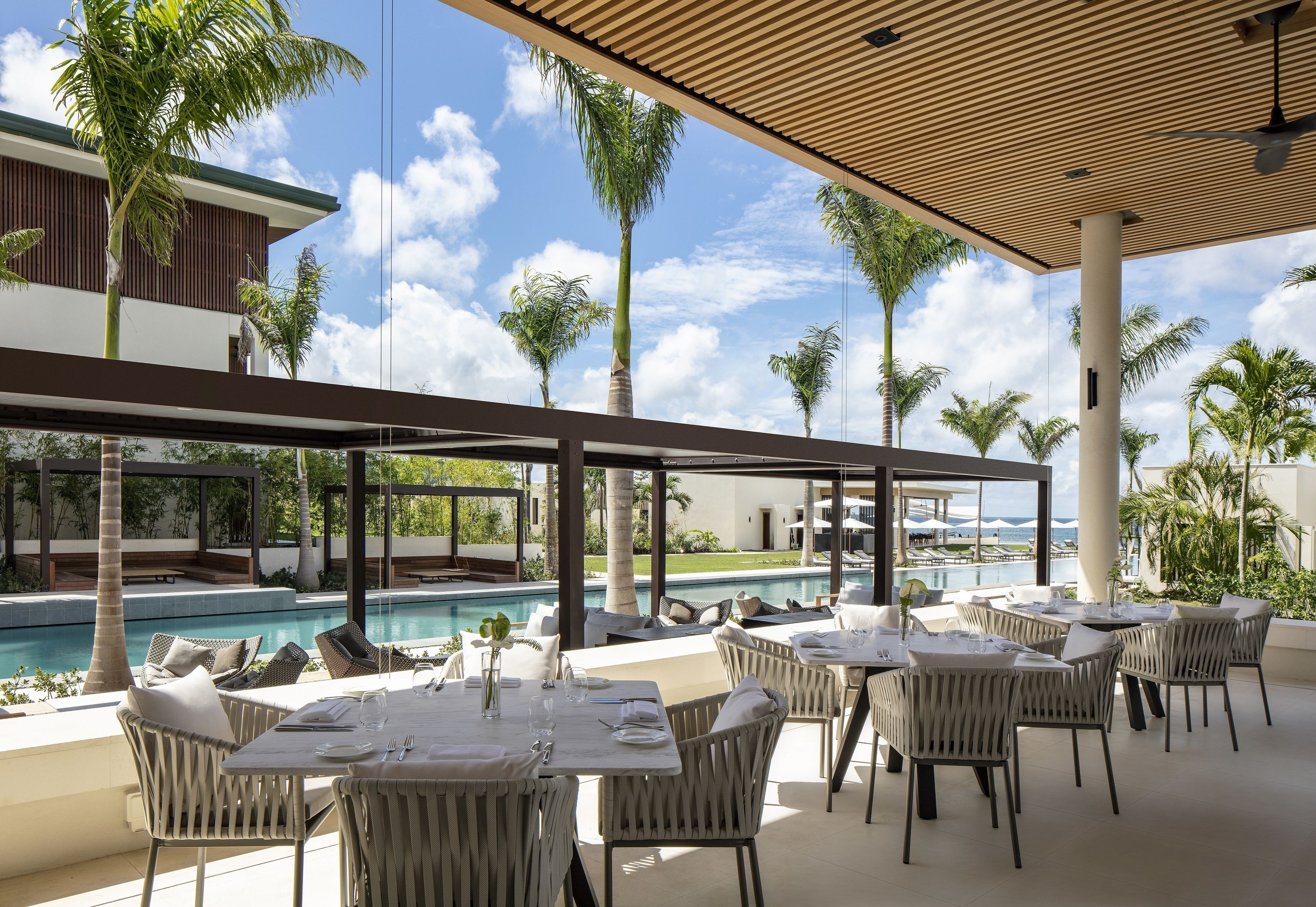 Un área de comedor al aire libre con mesas y sillas, con vista a una piscina y palmeras.