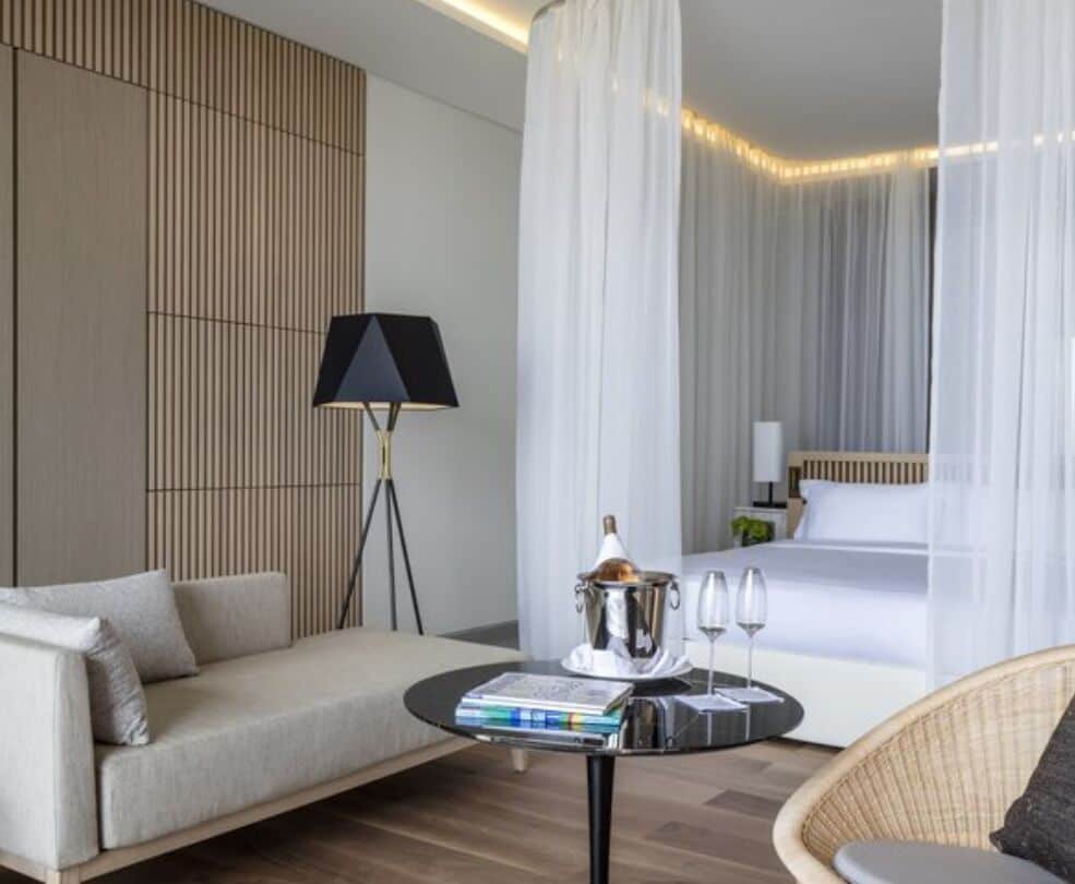A modern hotel room with a sofa, a round table with books and glasses, a lamp, and a bed with a canopy.