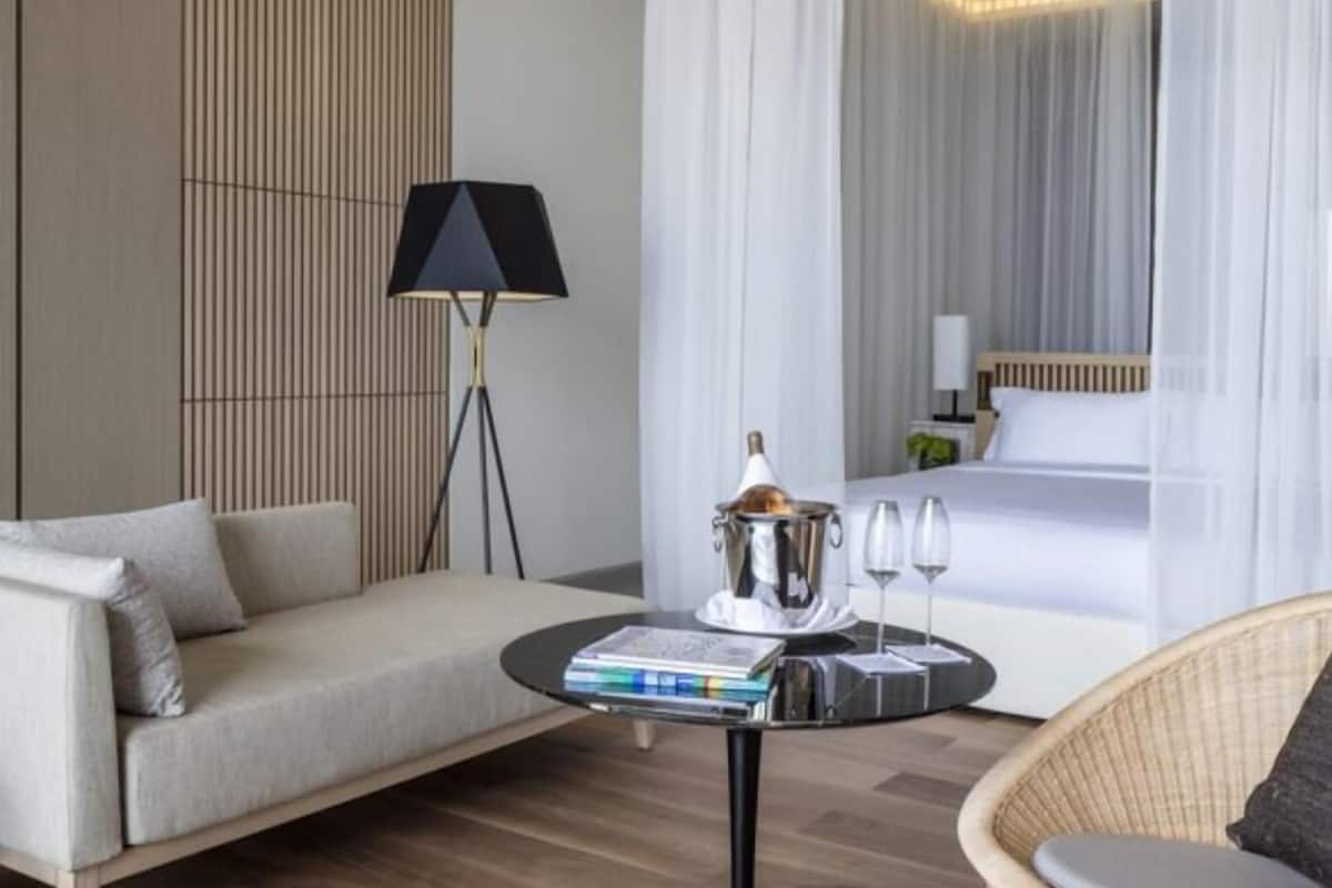 A modern hotel room with a sofa, a round table with books and glasses, a lamp, and a bed with a canopy.
