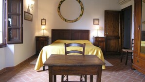 A bedroom with a bed, a wooden table, a chair, a round mirror on the wall, and a window with a curtain. - Tenuta Monti (Borgo San Lorenzo)