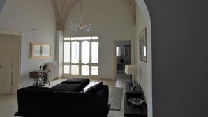 A spacious living room with a high arched ceiling, a chandelier, and a large window allowing natural light. - La Casa Del Mercante (Racale)