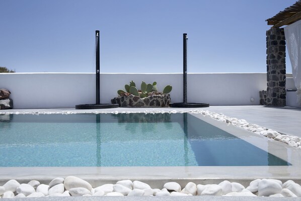 A pool with clear blue water, surrounded by white pebbles, with a cactus plant and two tall black poles in the background.