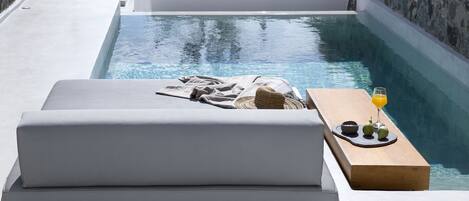 A modern poolside area with a lounge chair, a wooden table with a drink and fruits, and a stone wall backdrop.