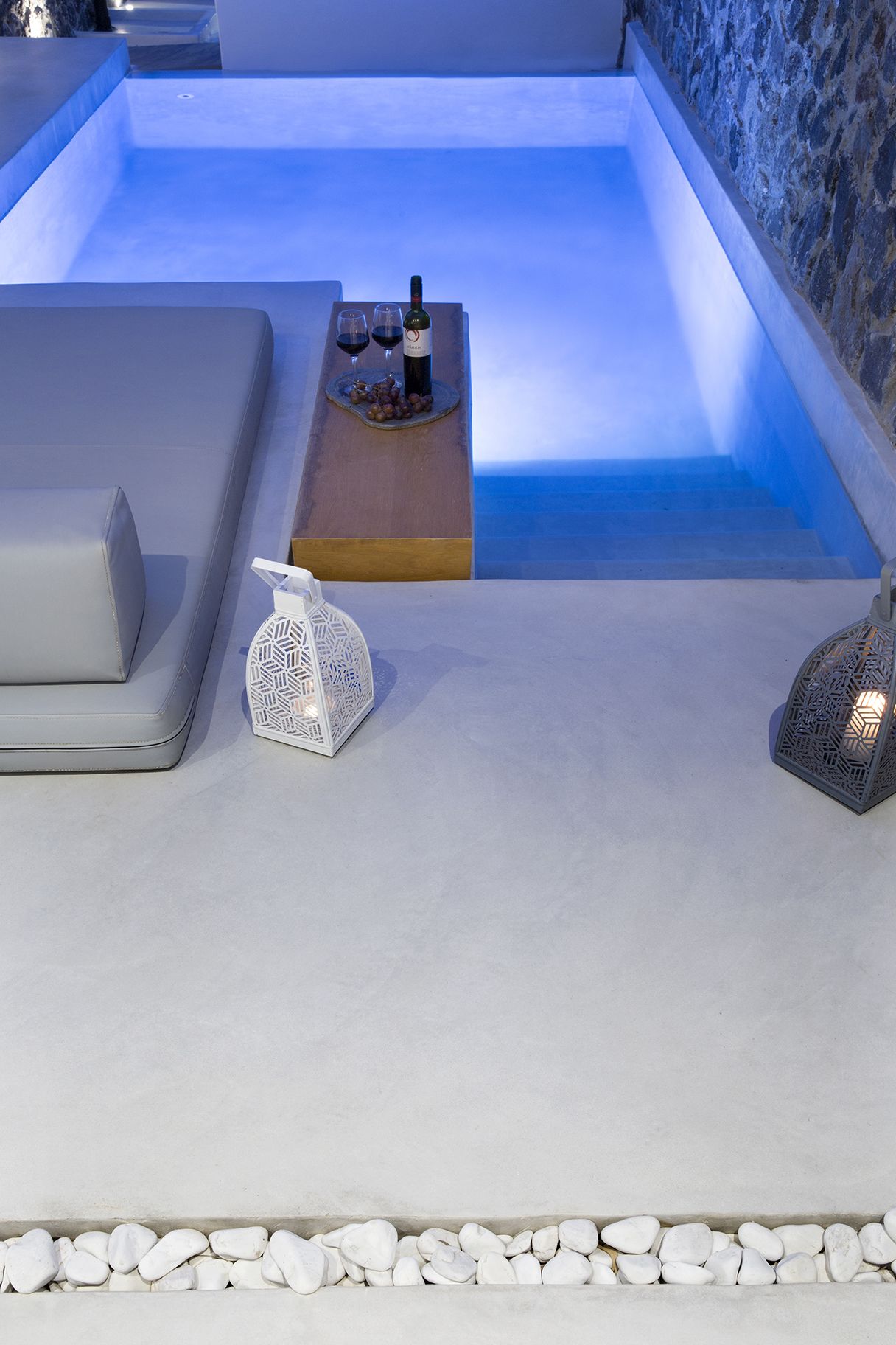 A modern outdoor relaxation area with a stone wall, a heated pool, and a seating area with a table set with wine glasses and a bottle.