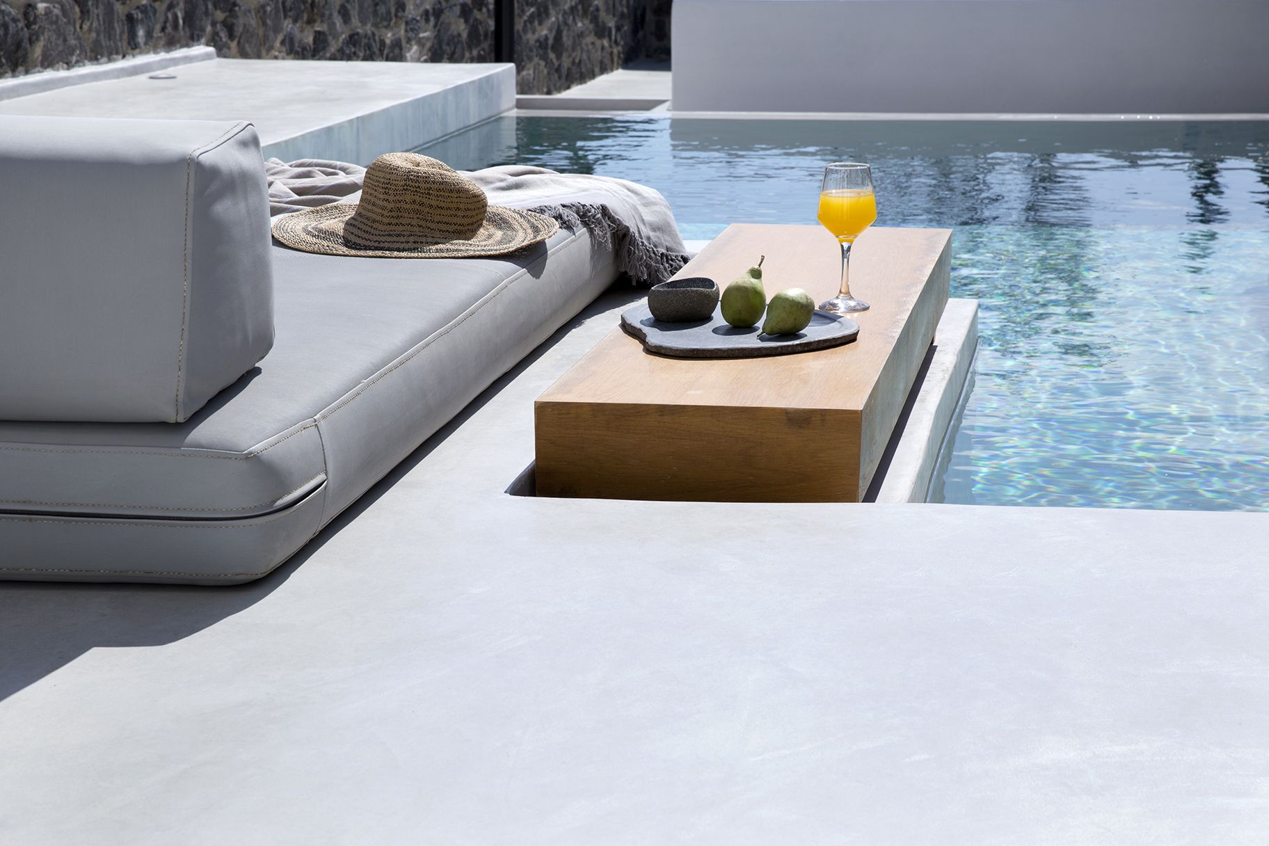 A poolside area with a white sofa, a wooden table with a glass of orange juice, a straw hat, and a bowl of fruit.