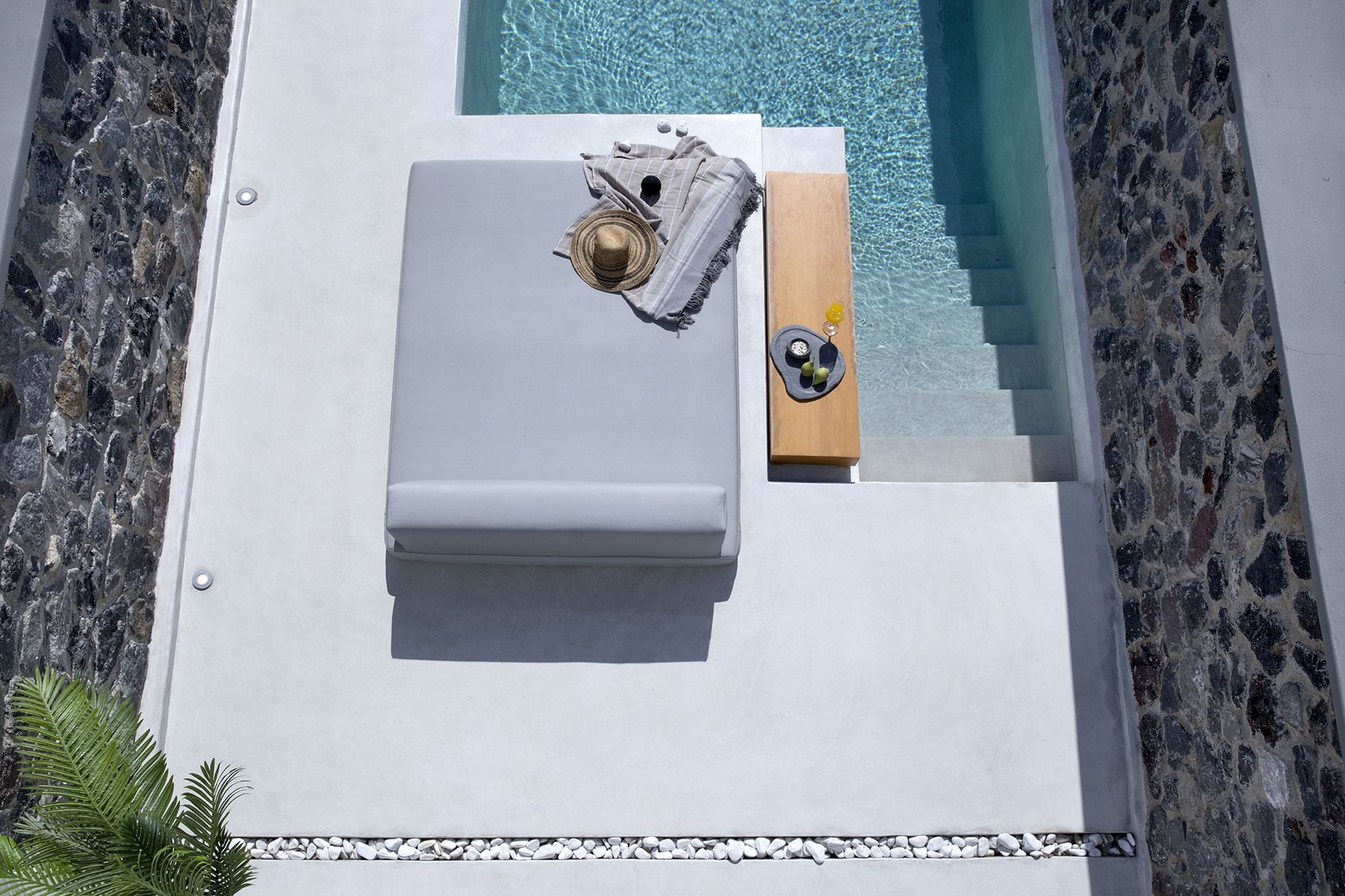 An aerial view of a poolside lounge area with a white lounge chair, a wooden side table with a drink tray, and a stone wall.