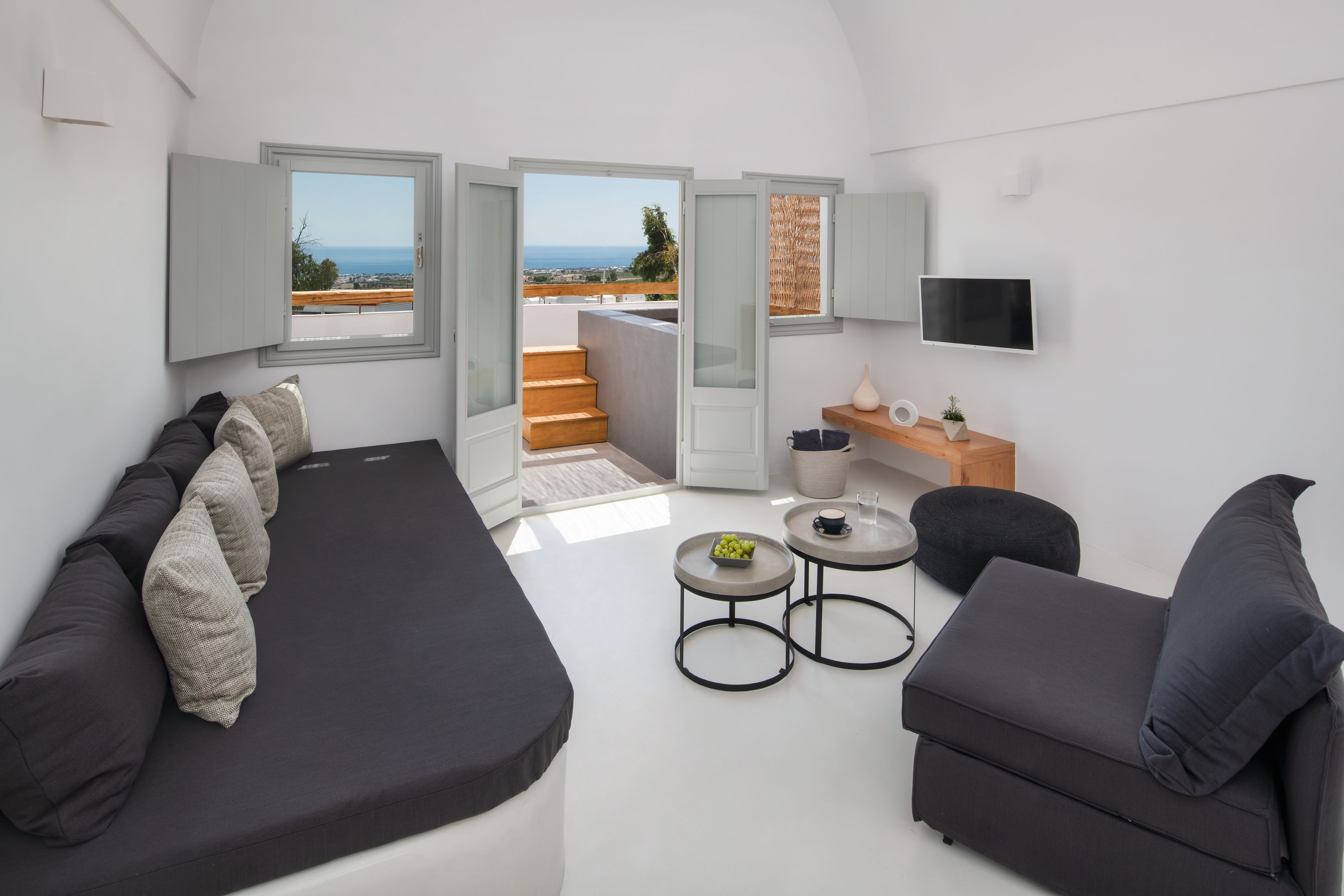 Un salón moderno con un sofá, dos sillones, una mesa de centro y una televisión. Hay dos ventanas con vistas al mar y un balcón.