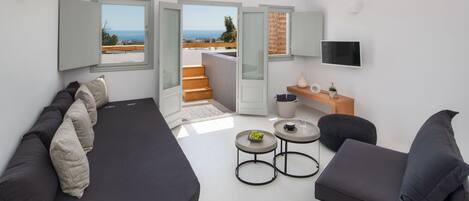 Un salón moderno con un sofá, dos sillones, una mesa de centro y una televisión. Hay dos ventanas con vistas al mar y un balcón.