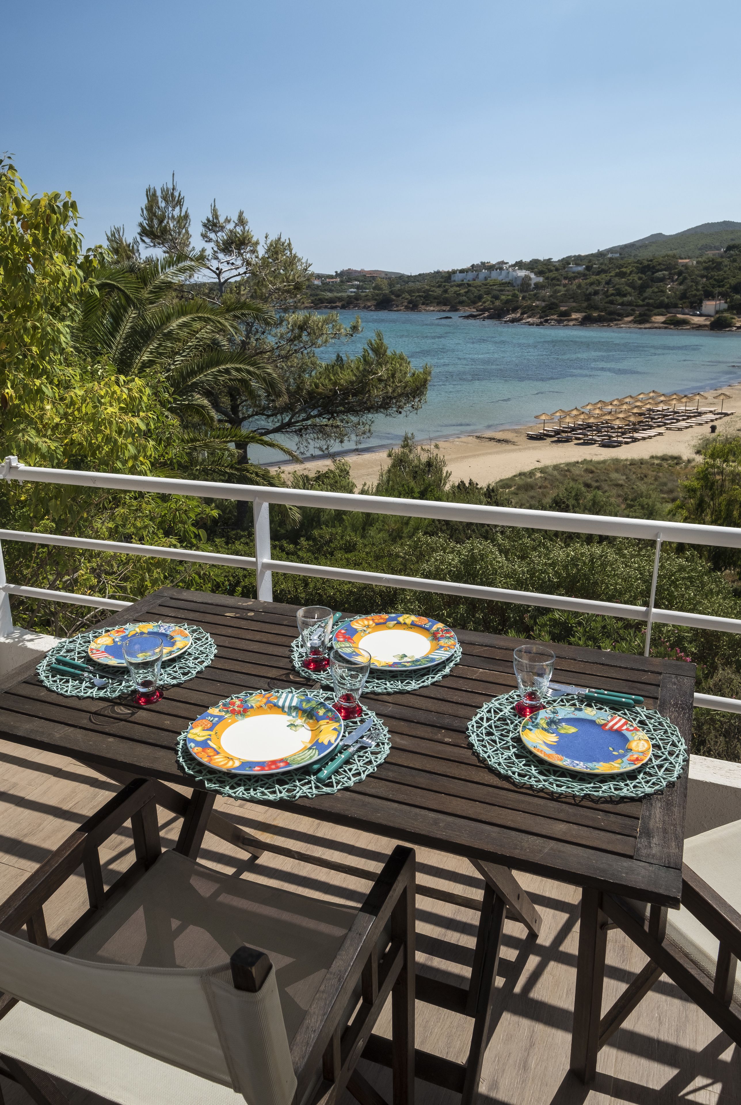 A wooden table with colorful plates and glasses, set on a balcony overlooking a beach and sea.
