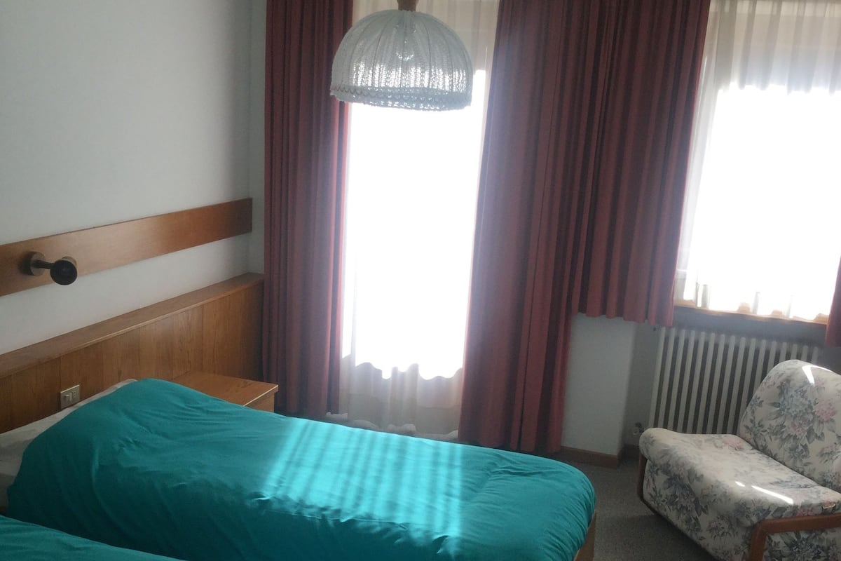 A hotel room with two beds, a chair, a radiator, and a window with curtains.