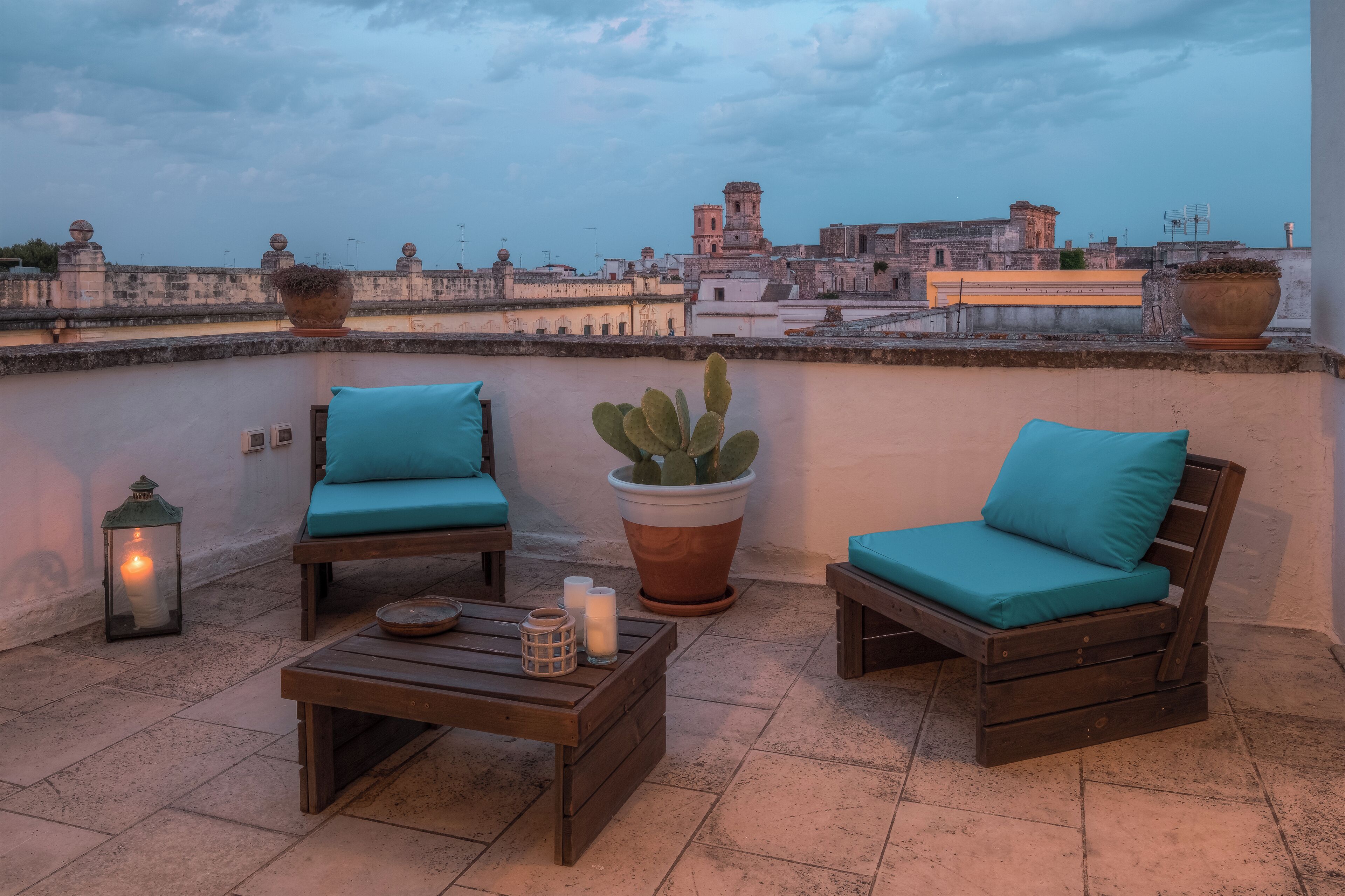 A rooftop terrace with two wooden chairs with teal cushions, a wooden coffee table, a potted cactus, and a lit lantern.