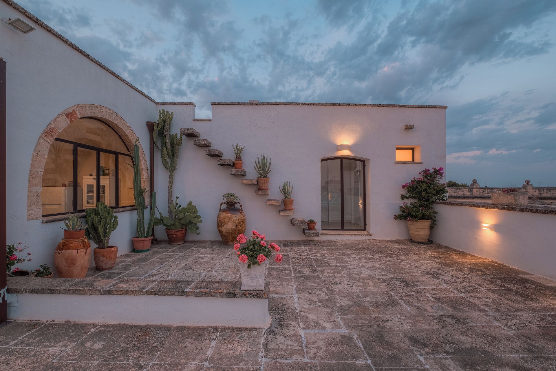 Una terraza en la azotea con suelo de piedra, plantas en macetas, un cactus y una puerta con panel de vidrio.