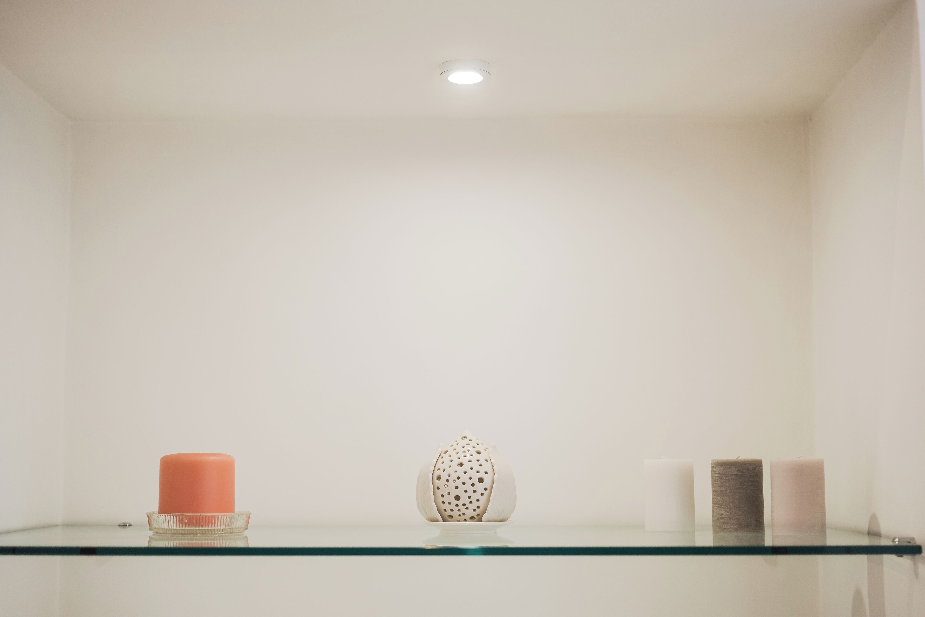 A minimalist room with a glass shelf displaying a candle, a decorative object, and two candles.