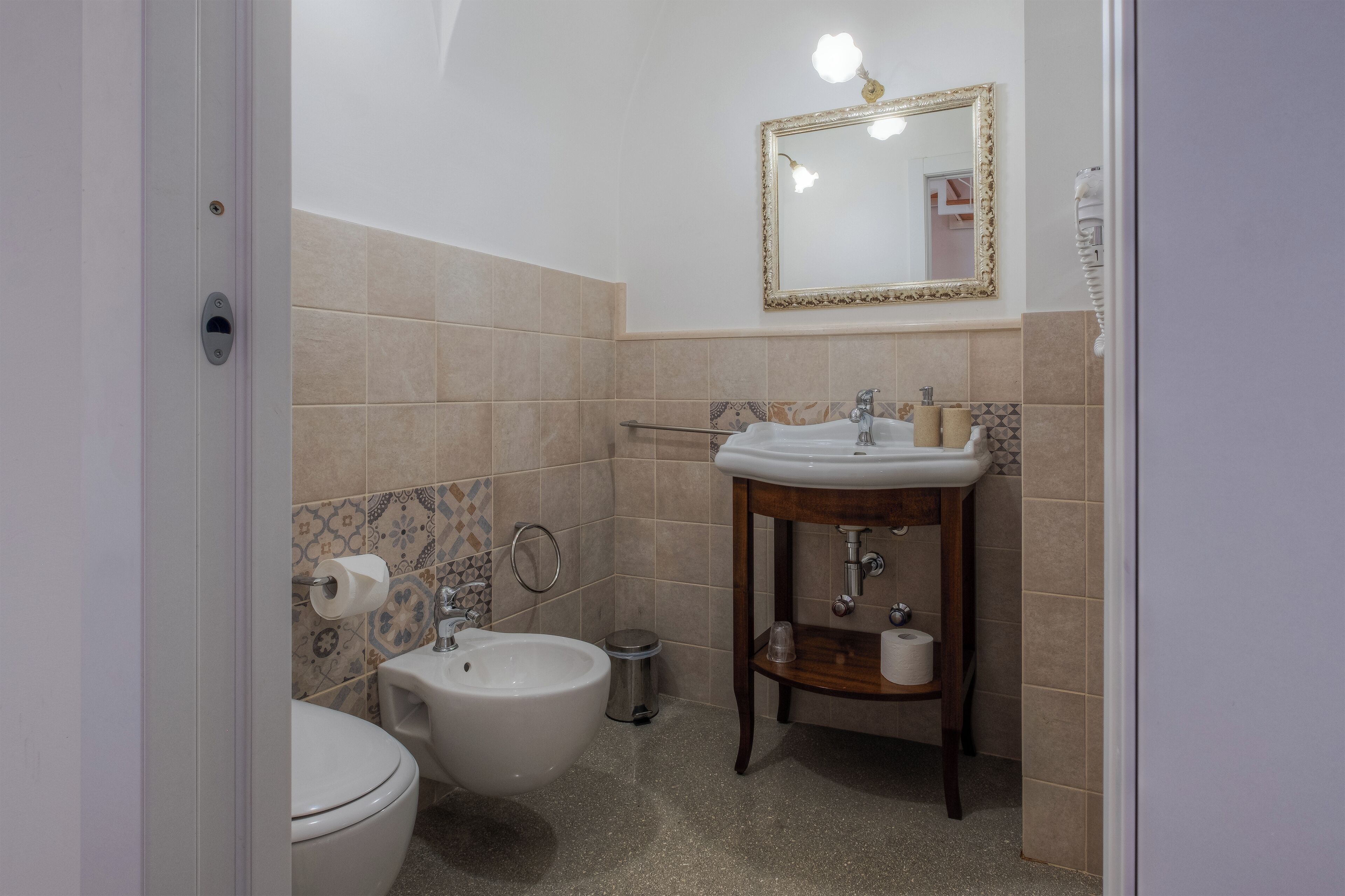 Casa de banho com sanita, bidé, lavatório e espelho com moldura decorativa.