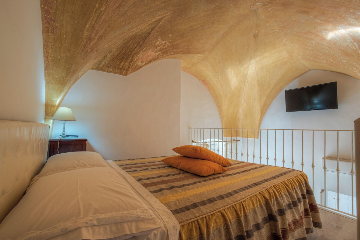 A bedroom with a vaulted ceiling, a bed with a striped bedspread, two pillows, a nightstand with a lamp, and a flat-screen TV mounted on the wall.