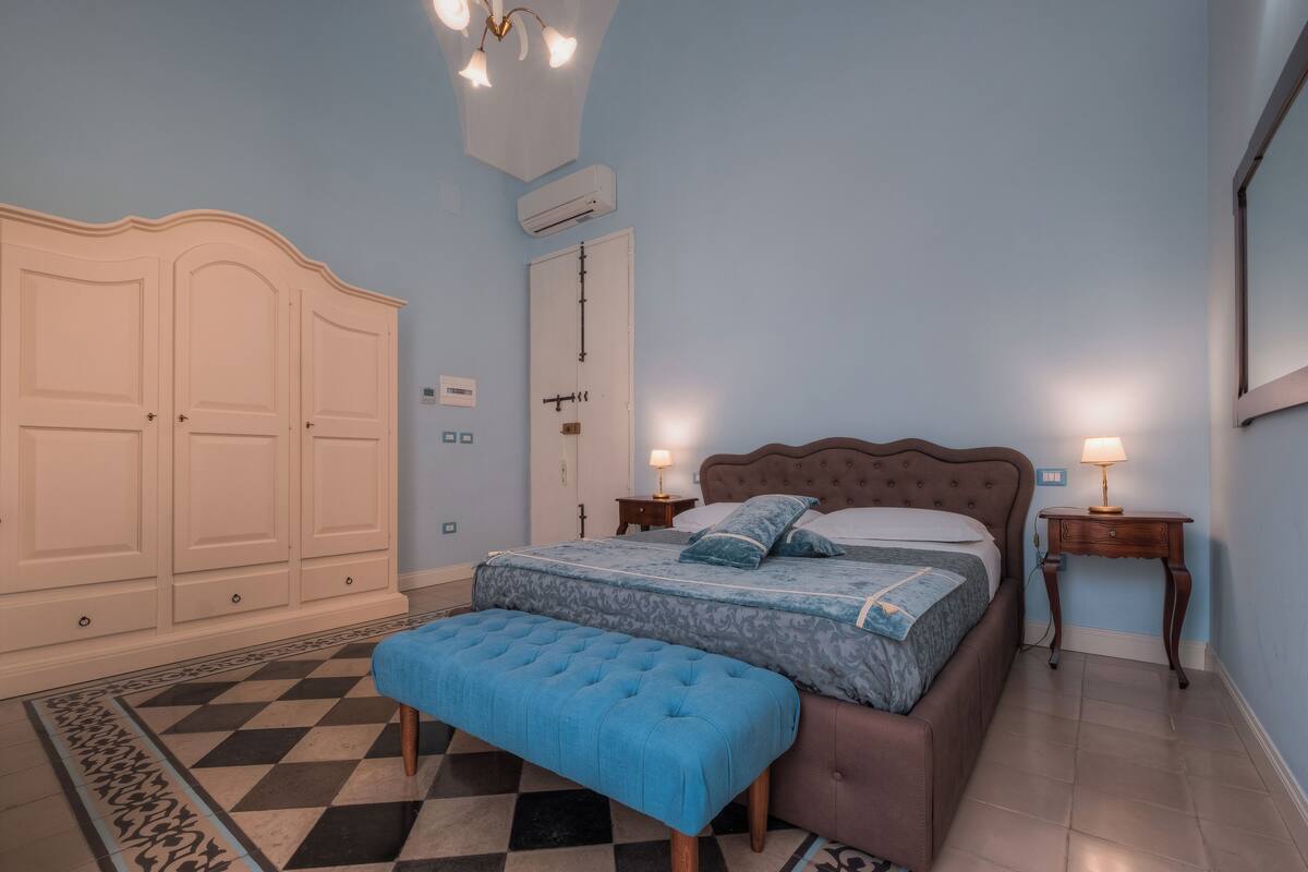 Une chambre à coucher comprenant un lit, un banc, une armoire, une table de chevet et un climatiseur fixé au mur.