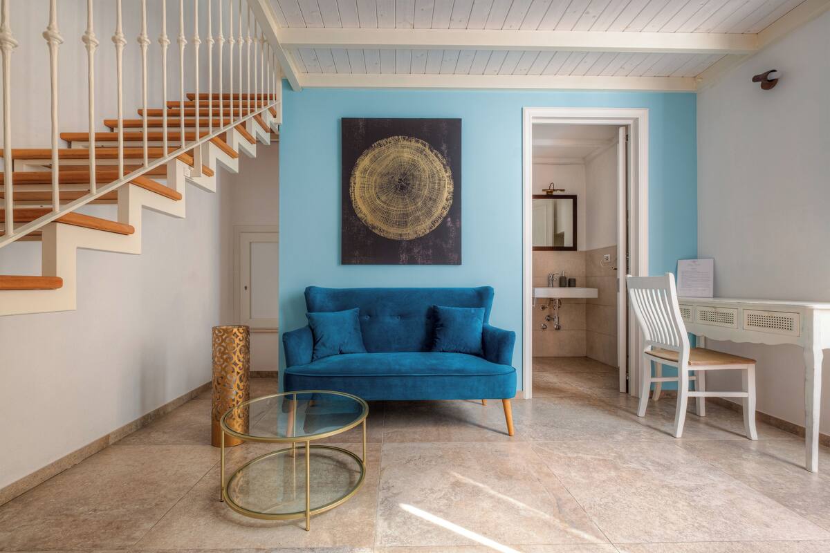 Un salon avec un canapé bleu, une table basse en verre, un escalier en bois et un coin repas comprenant une table et des chaises blanches.