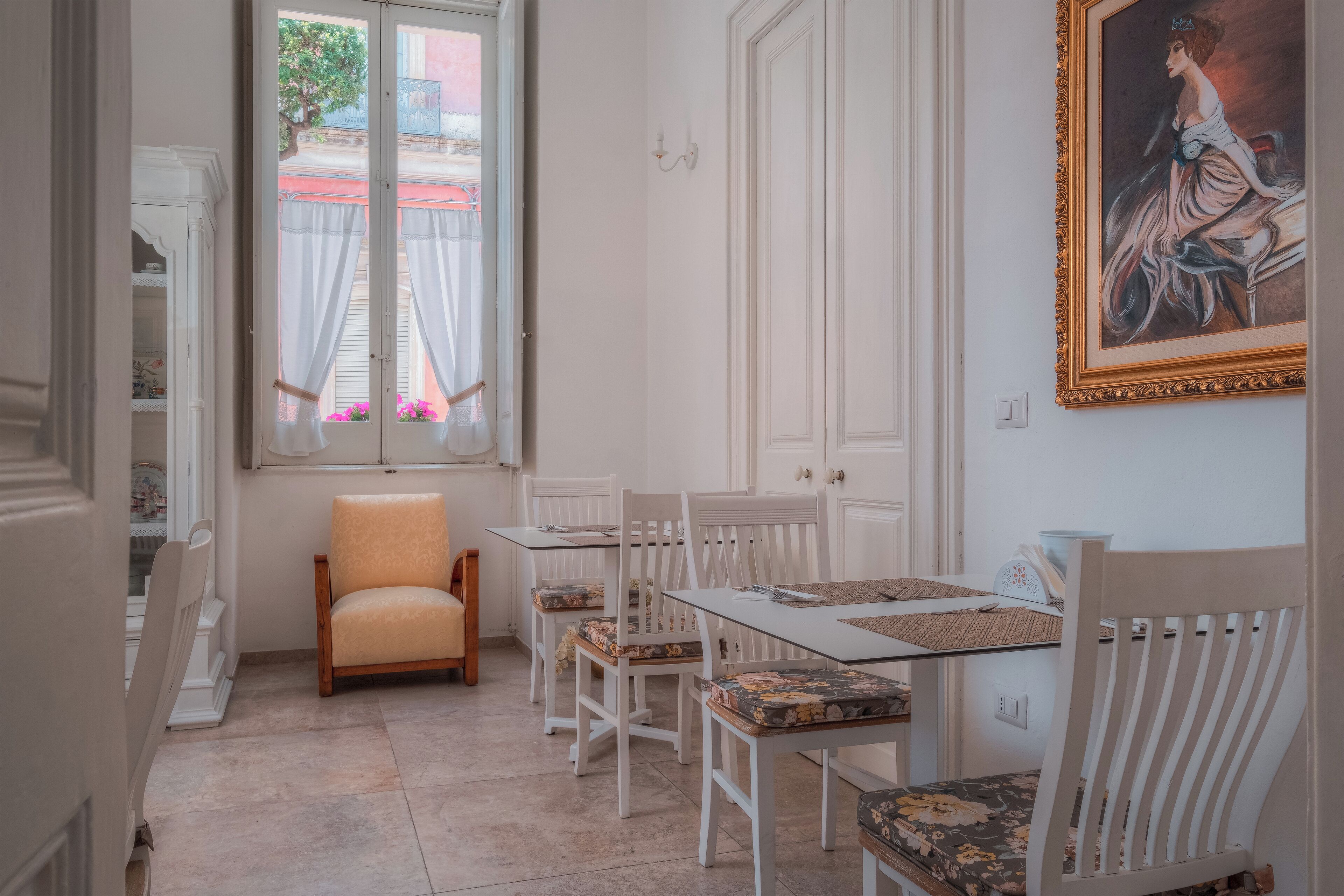 A dining area with white furniture, a window with curtains, and a framed painting on the wall.