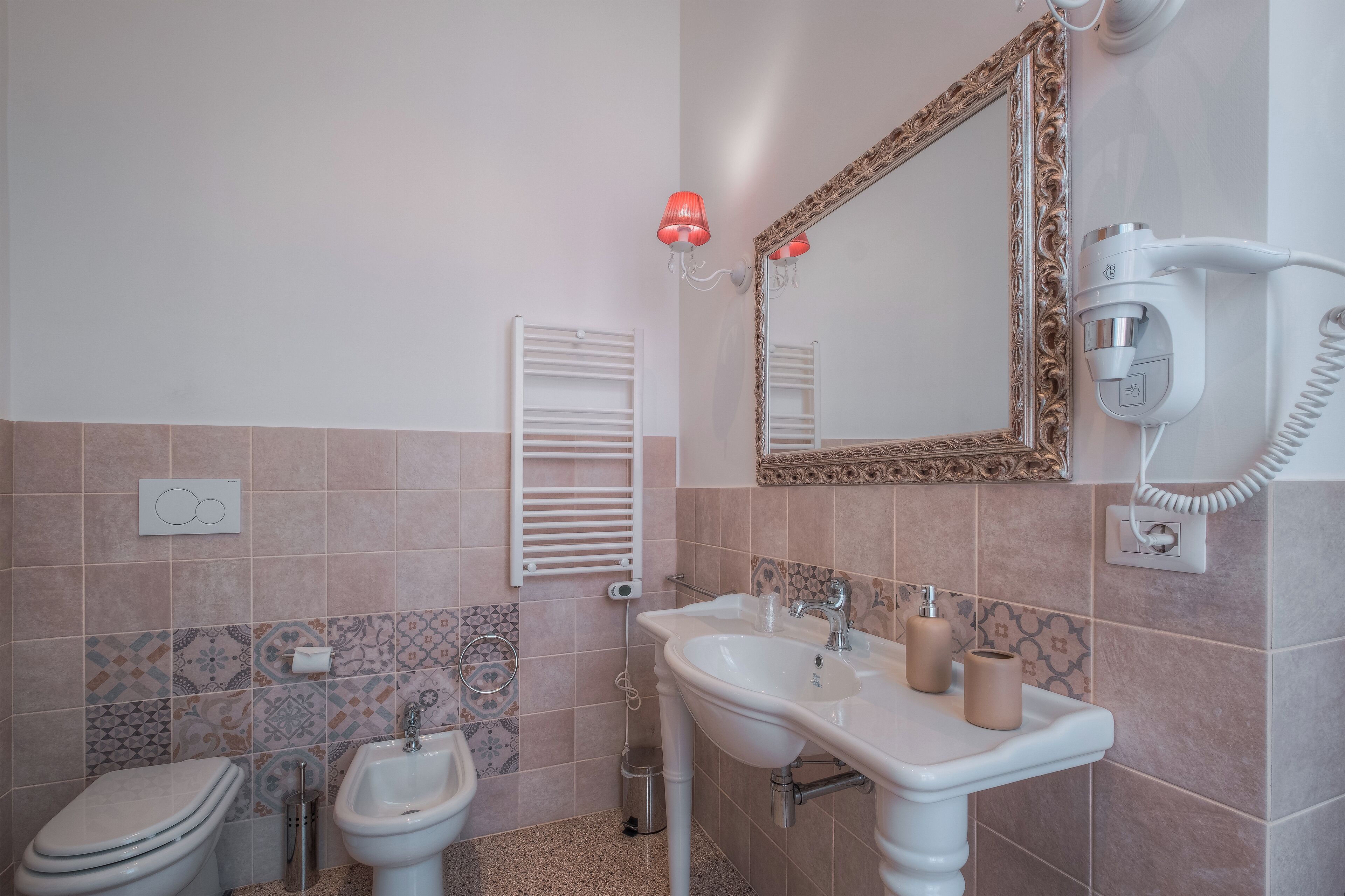 A bathroom with a sink, bidet, and toilet, a mirror with an ornate frame, and a hairdryer mounted on the wall.