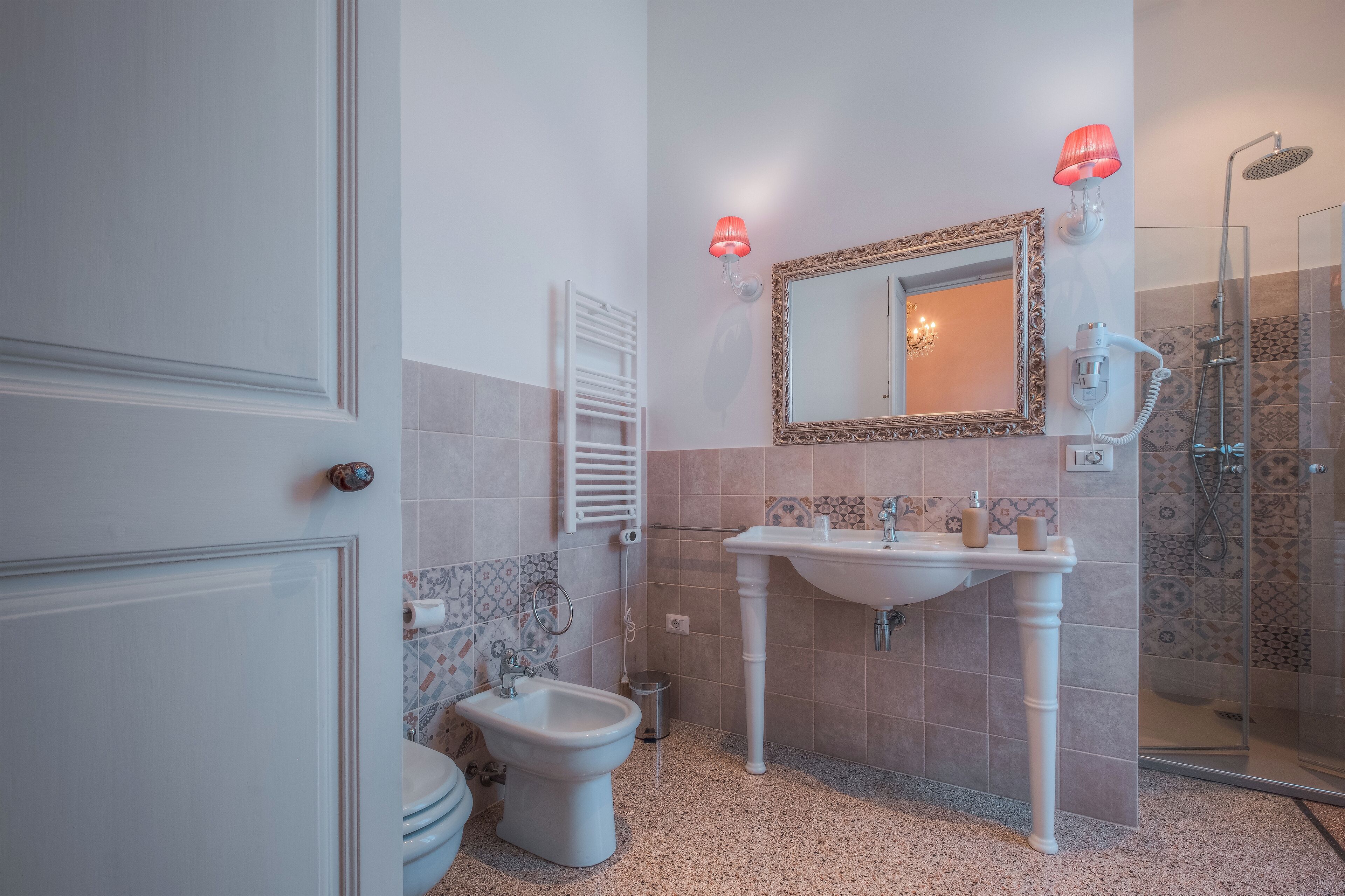 A bathroom with a pedestal sink, a bidet, a toilet, a shower, and a mirror with a decorative frame.