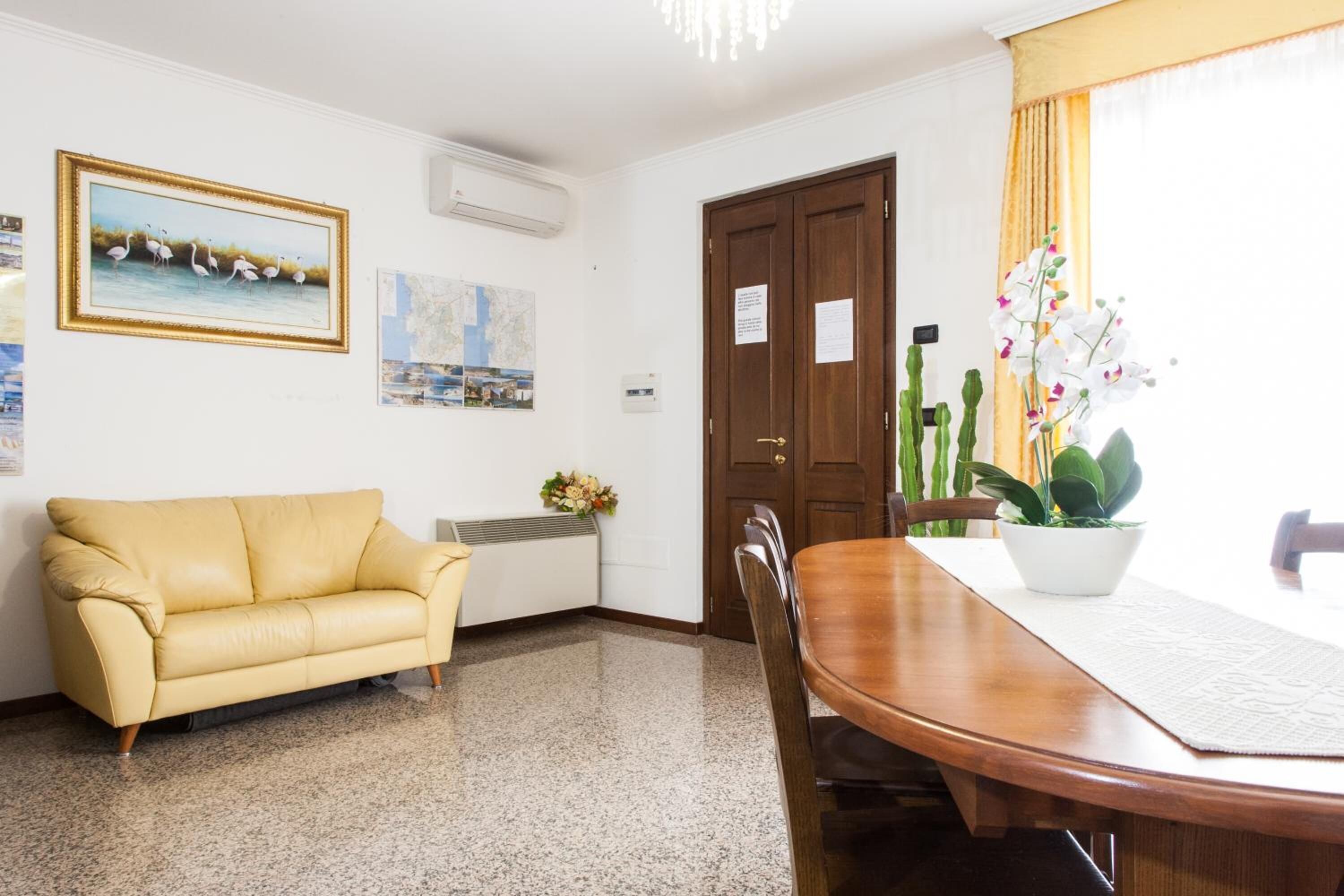 Une pièce comprenant une table à manger en bois, un canapé jaune, une chaise en bois, une plante en pot, un tableau encadré et une porte avec une pancarte.