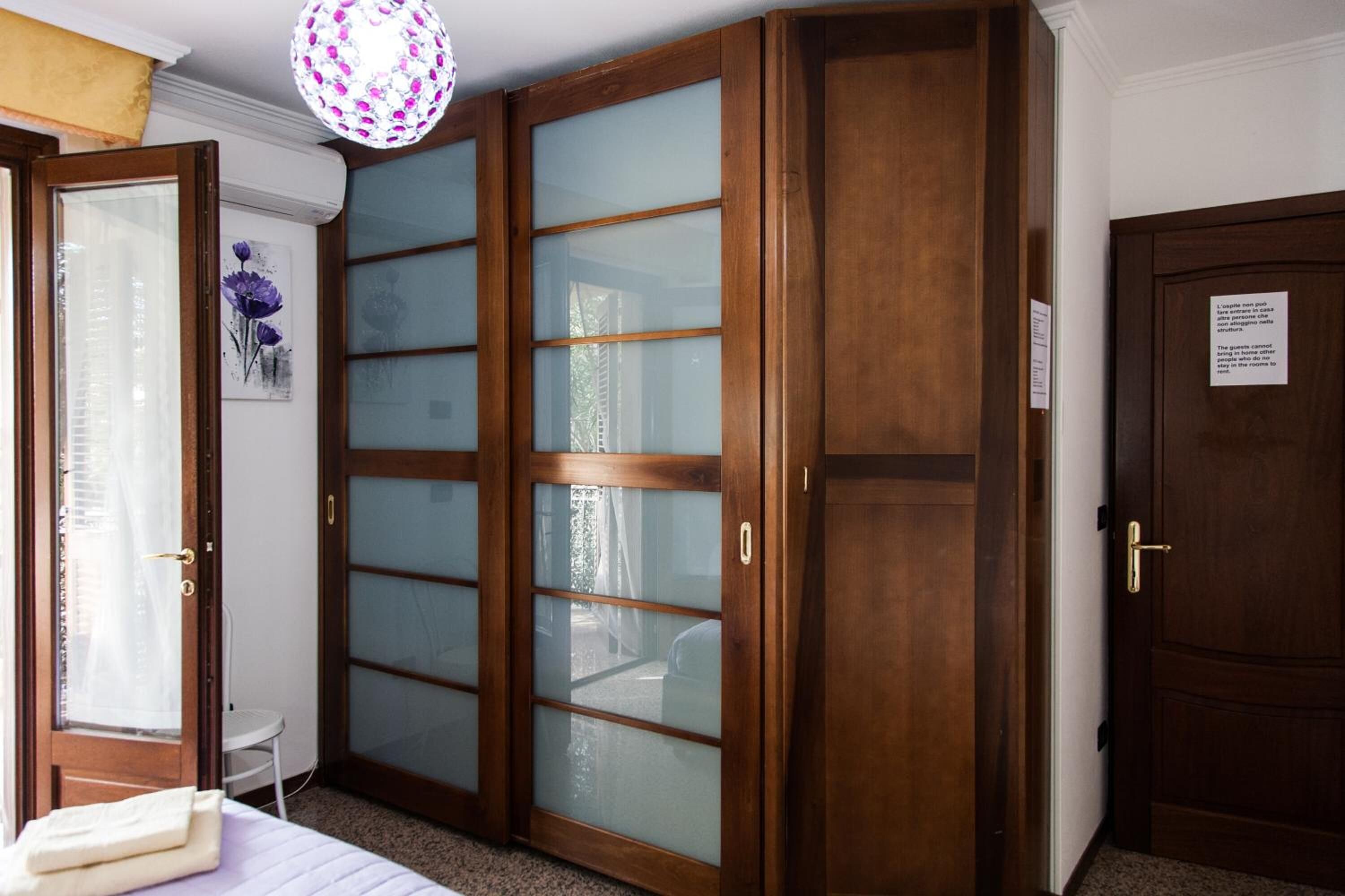Quarto com cama, guarda-roupa de madeira com portas de vidro jateado, uma mesinha branca e um abajur decorativo.
