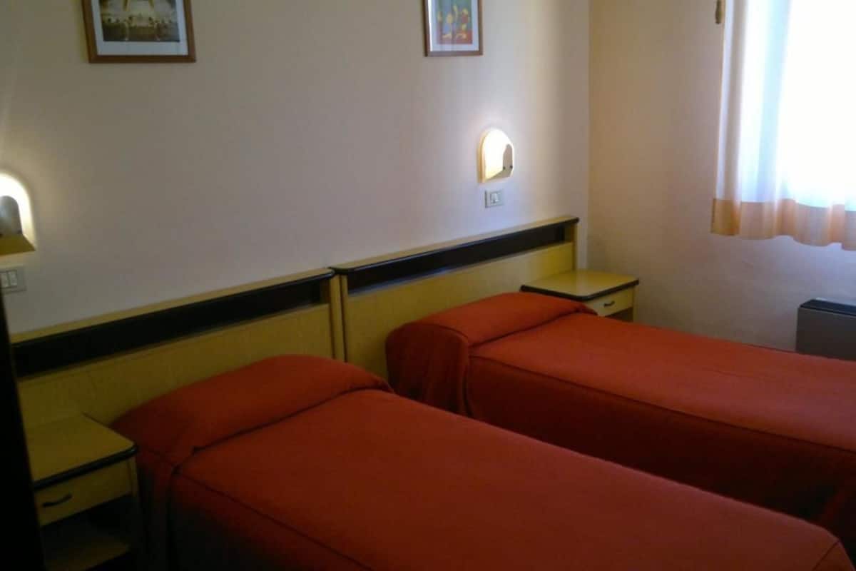 A hotel room with two single beds, a bedside table, a nightstand, a lamp, and a window with curtains.