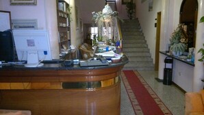 A reception area with a wooden desk, computer, and various items on the counter. A staircase is visible in the background. - Hotel Centrale (Argenta)