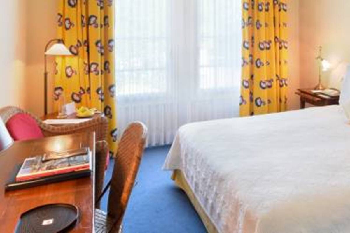 A hotel room with a bed, a desk with a chair, a lamp, and curtains with a cartoon pattern.