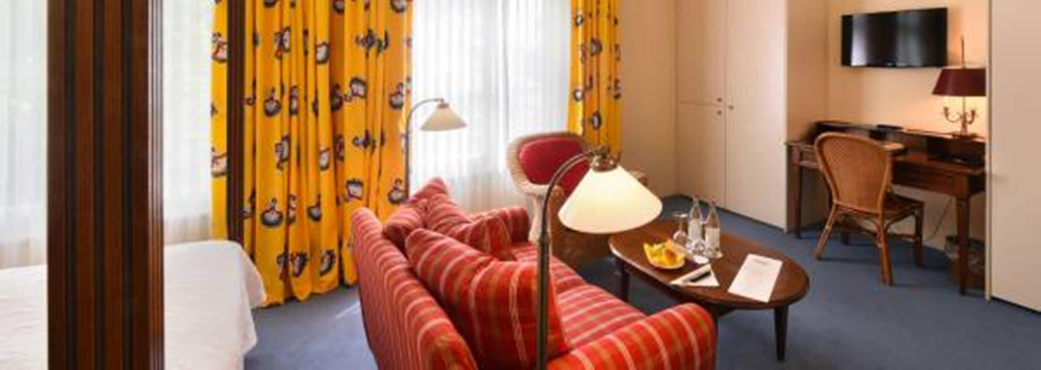 Een hotelkamer met een rood gestreepte bank, een kleine tafel met een lamp, een bureau met een stoel en geel gestreepte gordijnen.