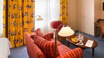 Een hotelkamer met een rood gestreepte bank, een kleine tafel met een lamp, een bureau met een stoel en geel gestreepte gordijnen.