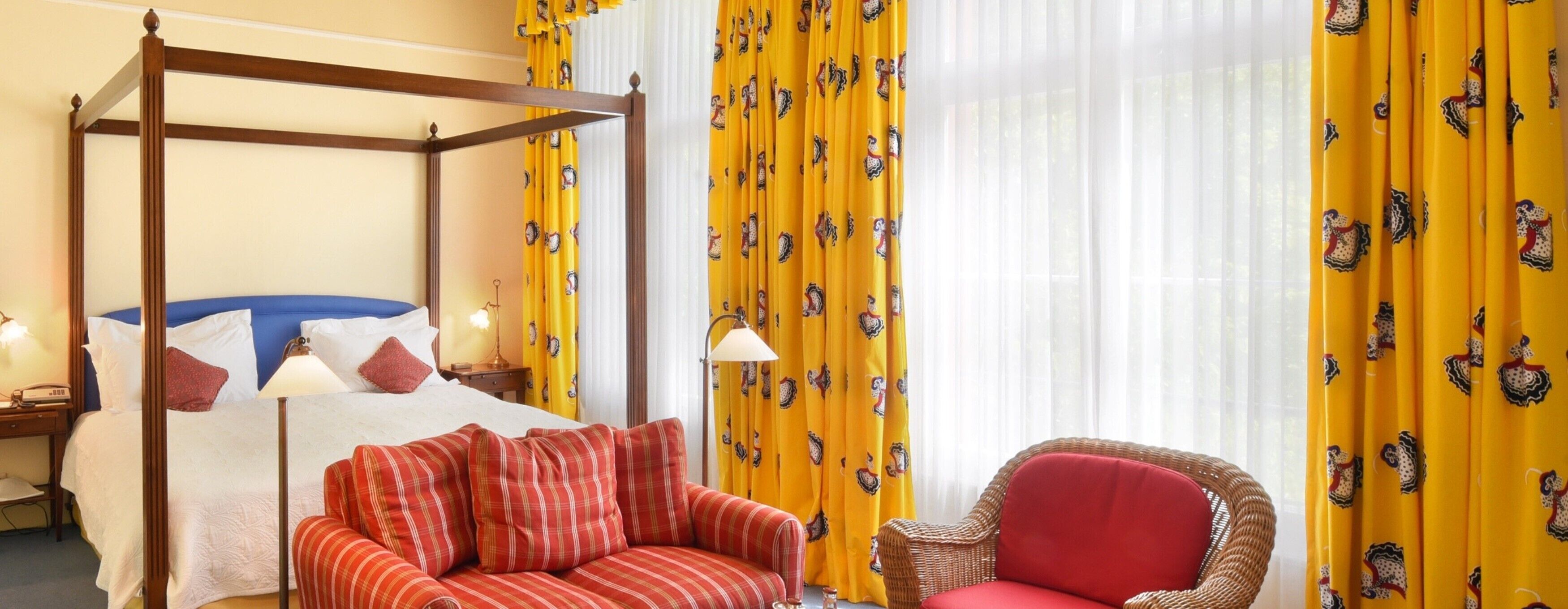 A hotel room with a four-poster bed, a red sofa, a wicker chair, and yellow curtains with a pattern.