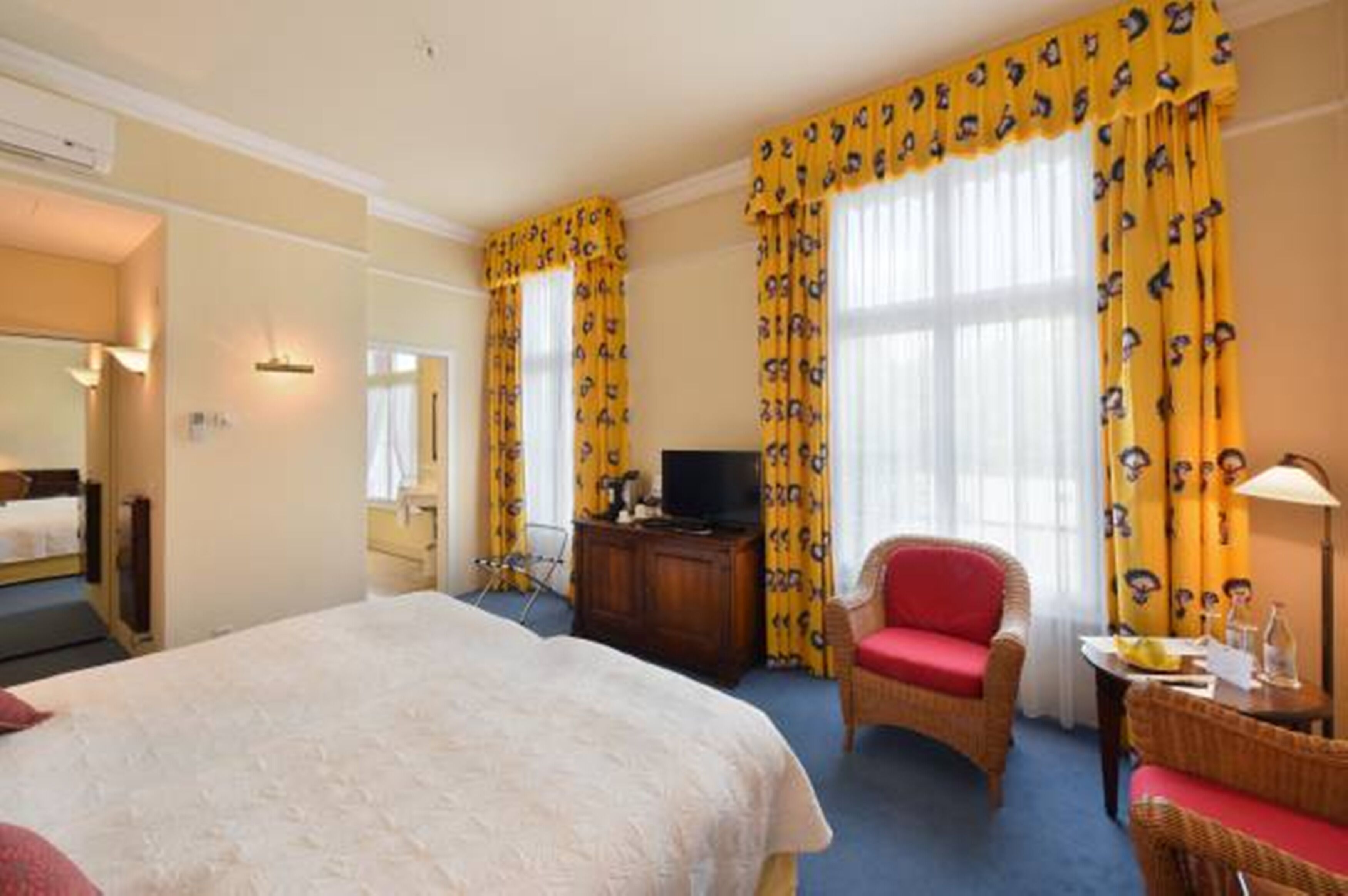 Habitación de hotel con una cama grande, un televisor sobre un mueble de madera, una silla roja y cortinas amarillas con estampado.