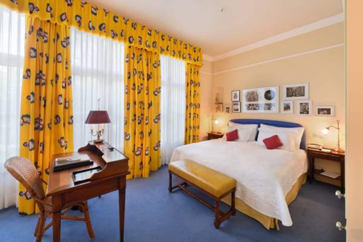 A hotel room with a large bed, a desk with a lamp, a chair, and a bench. There are curtains with a pattern, a wall with framed pictures, and a nightstand with a lamp.