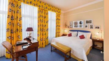 A hotel room with a large bed, a desk with a lamp, a chair, and a bench. There are curtains with a pattern, a wall with framed pictures, and a nightstand with a lamp.