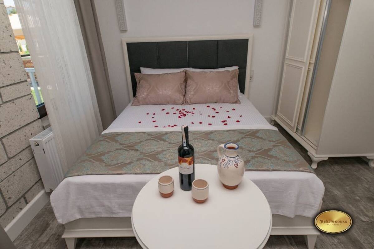 Un lit bien fait, recouvert d’une couvre-lit à motifs, avec deux oreillers, une bouteille de vin et deux verres posés sur une petite table ronde placée à côté du lit.