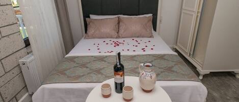Ein ordentlich bezogenes Bett mit einem gemusterten Überwurf, zwei Kissen sowie eine Weinflasche und zwei Gläser auf einem kleinen runden Tisch daneben.