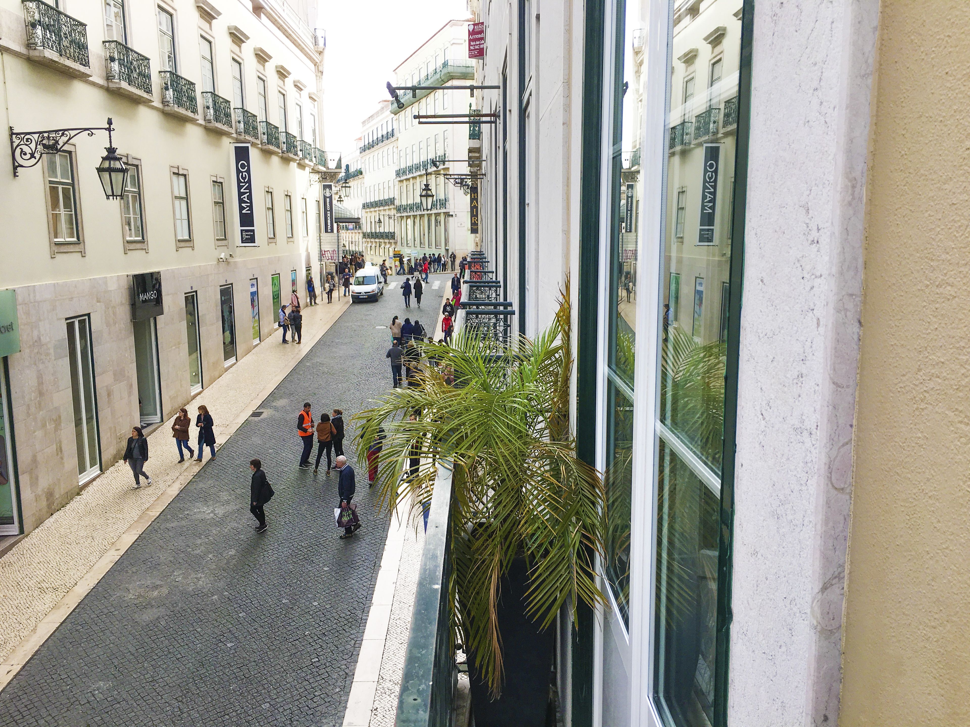 A cobblestone street lined with buildings, some with balconies and signage for businesses like "MANGO" and "HAIR".