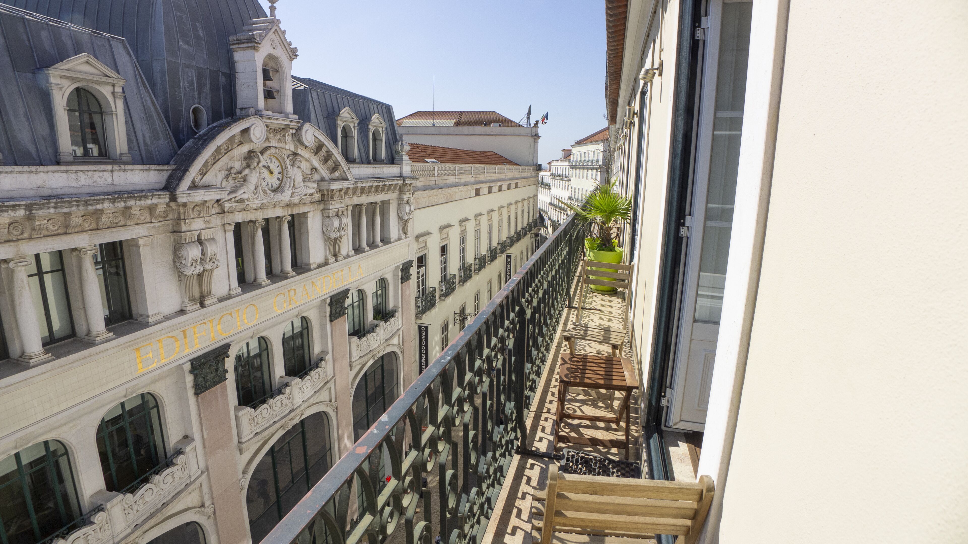 A narrow balcony with a wooden bench, overlooking a street with historic buildings and a clear sky.