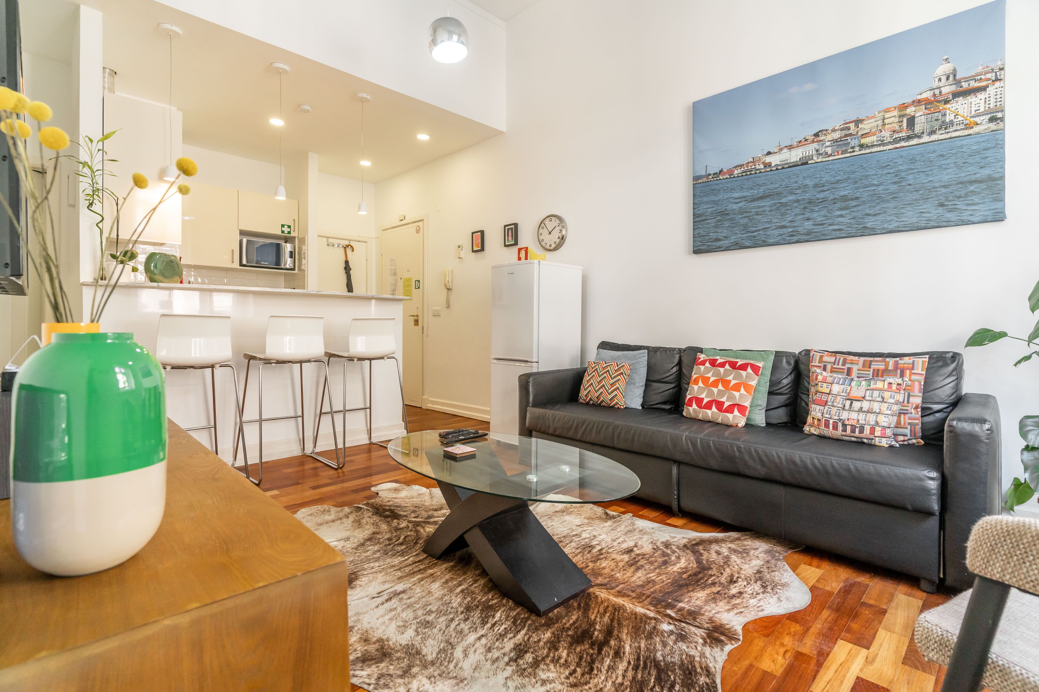 A modern living room with a glass coffee table, a leather sofa with patterned cushions, a dining area with white chairs, and a kitchen in the background.
