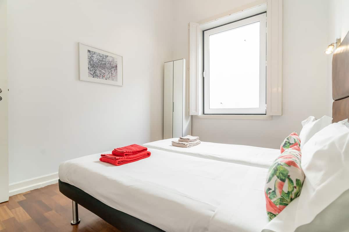 A hotel room with a bed, a window, a framed picture on the wall, and a bedside table with towels.