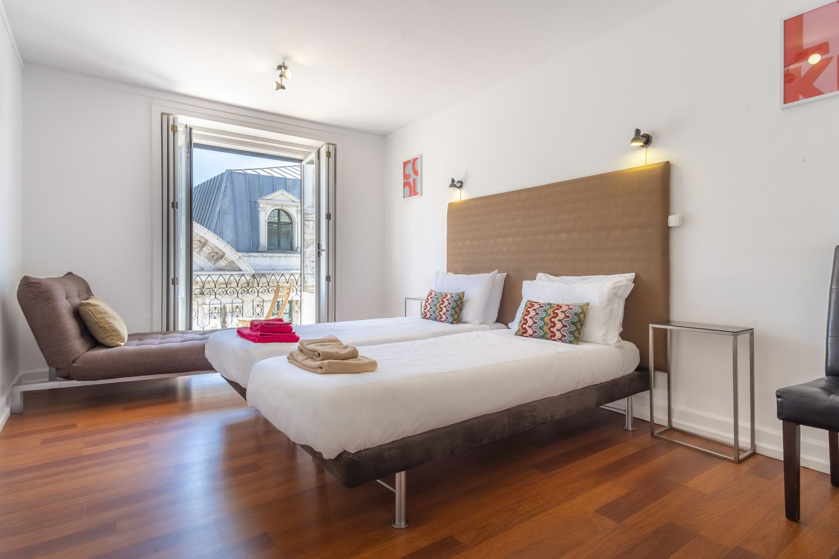 A hotel room with two beds, a sofa, a small table, and a view of a building through an open door.