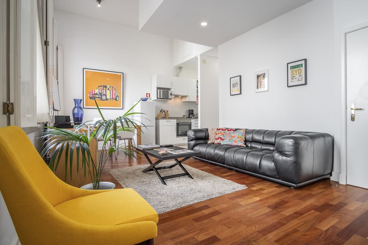 A modern living room with a yellow armchair, a black leather sofa, and a coffee table. There is a kitchen in the background with white cabinets and a microwave.